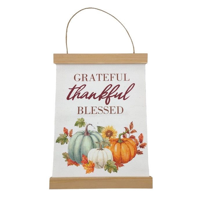 A hanging fabric wall sign with a light wood top and bottom border, featuring the phrase 'Grateful thankful blessed' above an autumn illustration of pumpkins, sunflowers, and leaves. A thin rope hanger is attached at the top.