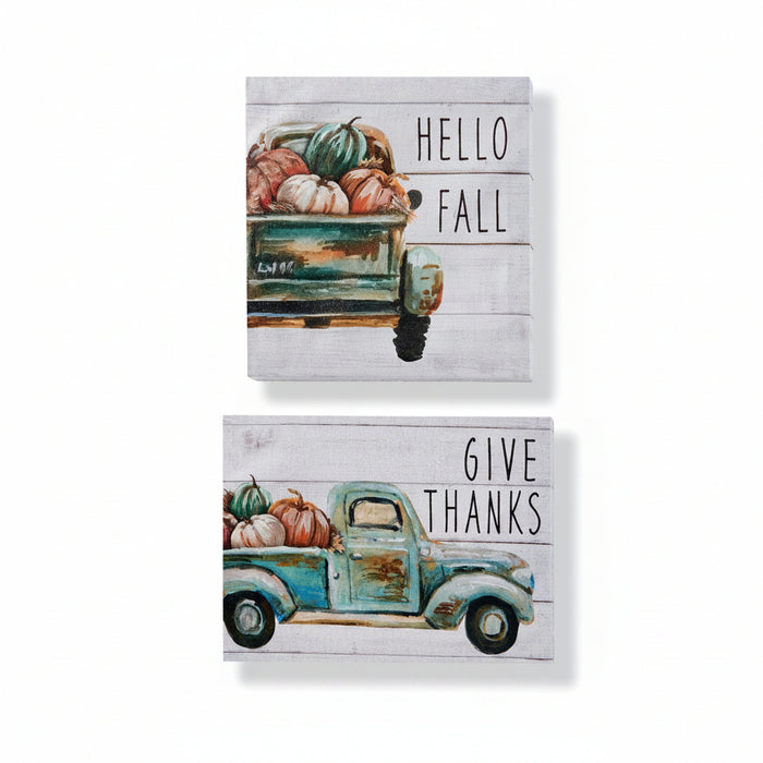 Two square canvas wall art pieces with a rustic fall theme. The top canvas reads 'HELLO FALL' and shows a teal pickup truck loaded with pumpkins on a whitewashed wood background.