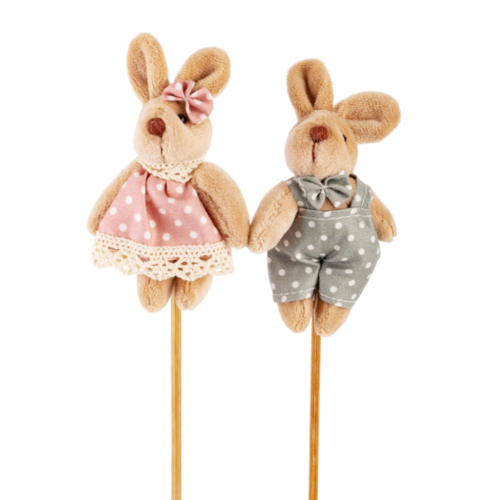 Two plush toy bunnies, one in a pink dress with a bow and the other in a gray outfit with a bow tie, on sticks against a white background.