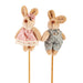 Two plush toy bunnies, one in a pink dress with a bow and the other in a gray outfit with a bow tie, on sticks against a white background.