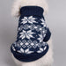 A white fluffy dog wearing a navy blue knit sweater with a white snowflake and diamond pattern, shown from behind against a neutral gray background.