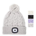 A light gray cable-knit pom-pom beanie with a built-in LED light on the front cuff, shown on a white background with four small color-option swatches on the right.