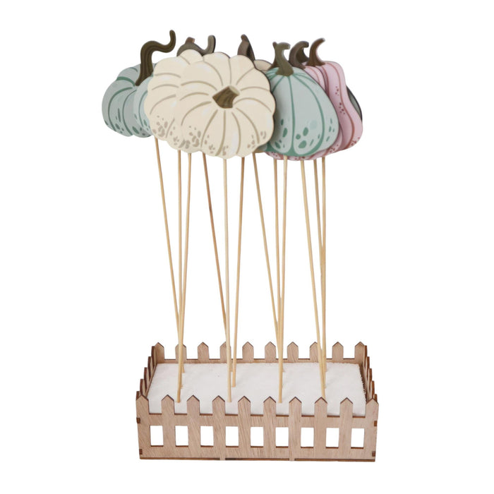 A decorative autumn-themed display featuring five hand-painted wooden pumpkins in pastel colors (cream, mint green, teal, and pink) with dark brown stems, mounted on wooden sticks over a small light wood picket fence base filled with white faux snow.