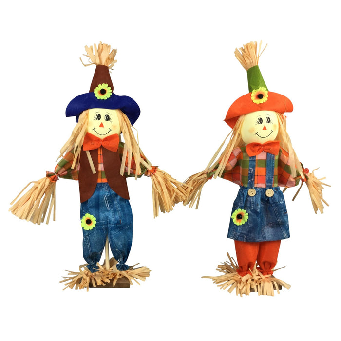 Two decorative scarecrows standing side by side on a plain white background. One scarecrow wears a blue hat, brown vest, and blue jeans with a yellow flower patch.