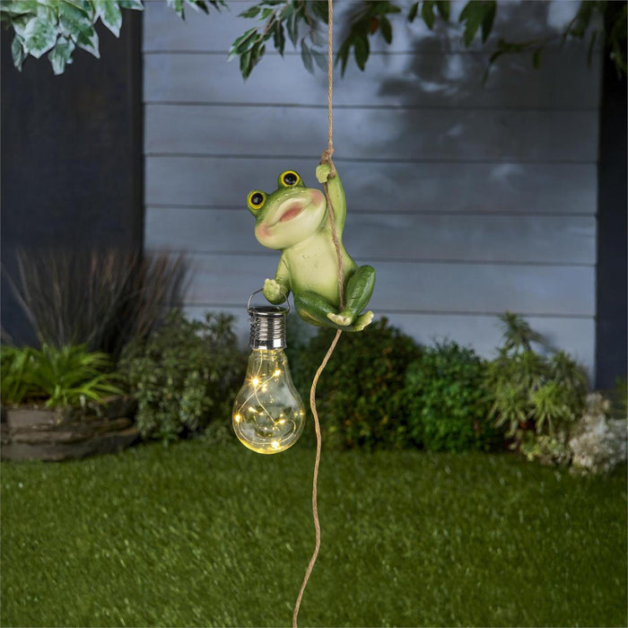 Frog-shaped light fixture hanging from a string with a light bulb, set against a garden background.
