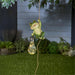 Frog-shaped light fixture hanging from a string with a light bulb, set against a garden background.