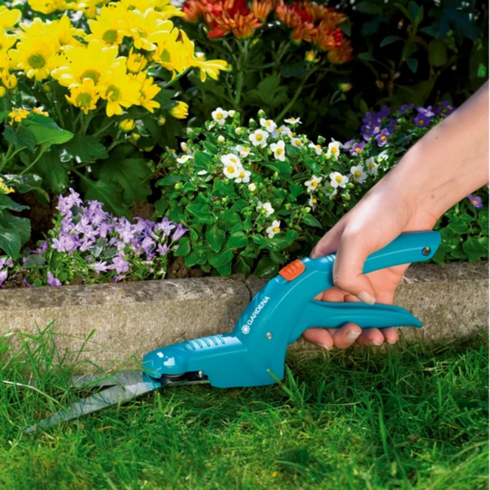 Person using a blue garden tool to trim grass near a flower bed with various flowers.