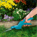Person using a blue garden tool to trim grass near a flower bed with various flowers.