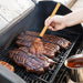 Person grilling steaks on a barbecue with a brush applying sauce.