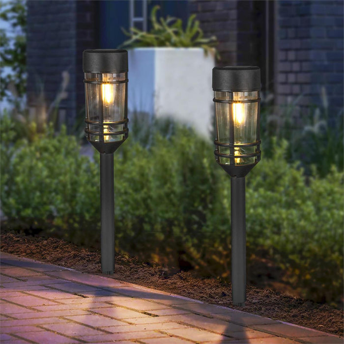 Two solar garden lights on stakes in a garden setting with a brick path.