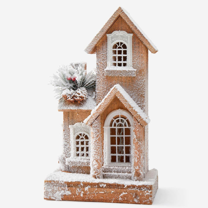 A decorative miniature house ornament with a rustic brown wood-grain finish and white faux snow dusting, featuring arched windows and a small snowy wreath accent on the side, set against a plain white background.