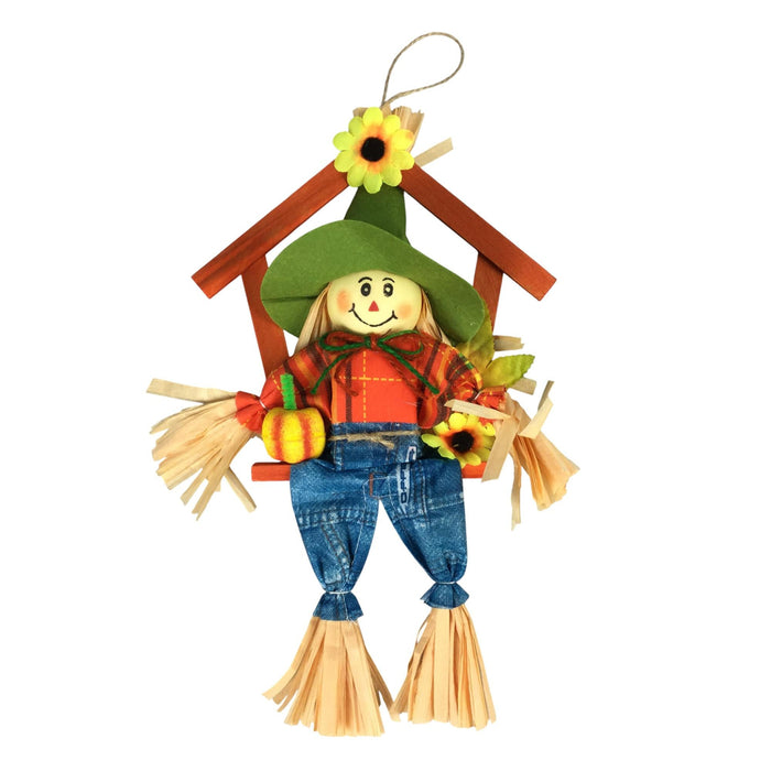A fabric scarecrow hanging decoration with a green hat, orange shirt, blue denim-style pants, straw hair and cuffs, a small pumpkin, sunflower, and wooden house frame on a white background.