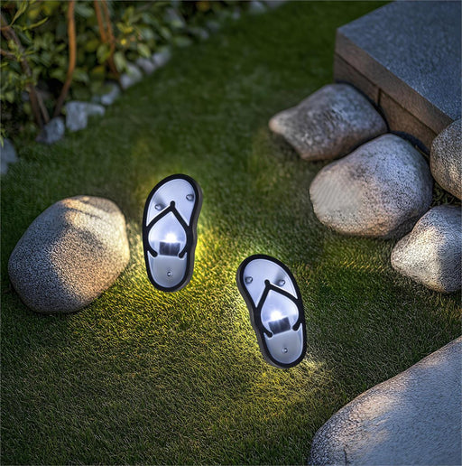 Two flip-flop-shaped solar lights on grass with rocks in the background