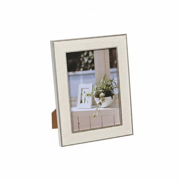 A vertical picture frame with a textured off-white/cream border and a silver-toned inner trim, shown standing on a small brown support against a plain white background. The frame contains a sample photo of a potted plant.