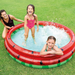 Two children splashing and playing inside a red and green inflatable watermelon-themed pool on a grassy lawn, while an adult supervises nearby.