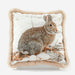 A square decorative throw pillow with a faux fur trim in a warm beige tone, featuring a printed or embroidered image of a brown and gray rabbit sitting in a snowy outdoor scene on the front.