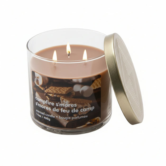 A three-wick scented candle in a clear glass jar with a metallic gold lid leaning against it, labeled 'campfire s'mores' with images of graham crackers and marshmallows on a white background.
