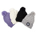 Four cable-knit pom-pom beanies in purple, cream, black, and light gray, arranged diagonally on a plain white background. The light gray beanie features a square black button with a white and blue eye motif on the cuff.