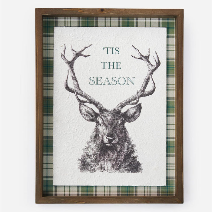 A framed holiday art print featuring a detailed black ink sketch of a stag with large antlers on a white textured background, surrounded by a green and white plaid border and a medium brown wooden frame.