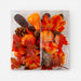 A square clear acrylic or glass decorative tray or block containing an autumn arrangement of faux pumpkins, red and orange maple leaves, and brown pinecones, photographed against a plain white background.