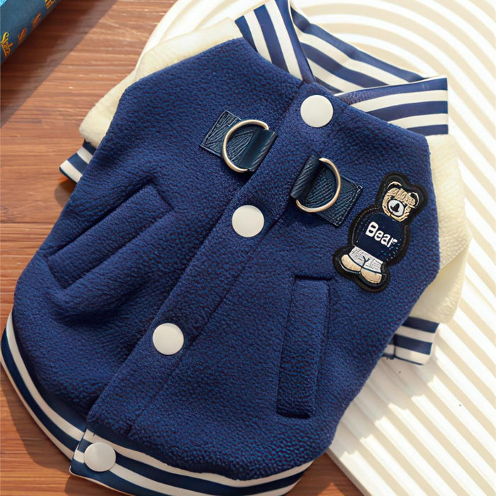 A navy blue fleece pet jacket with white buttons, striped navy and white collar and hem, and a bear patch on the chest, displayed on a striped plate over a wooden surface.