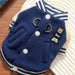 A navy blue fleece pet jacket with white buttons, striped navy and white collar and hem, and a bear patch on the chest, displayed on a striped plate over a wooden surface.