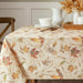 An autumn-themed tablecloth with a beige background and a pattern of colorful fall leaves and wheat stalks, styled on a dining table with white dishes and a white pitcher.