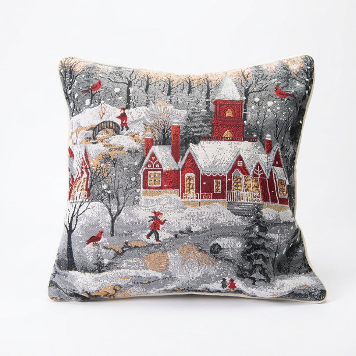 A square decorative throw pillow featuring a detailed winter village scene with red houses, snow-covered roofs, bare trees, and small figures in red clothing, shown against a plain white background.