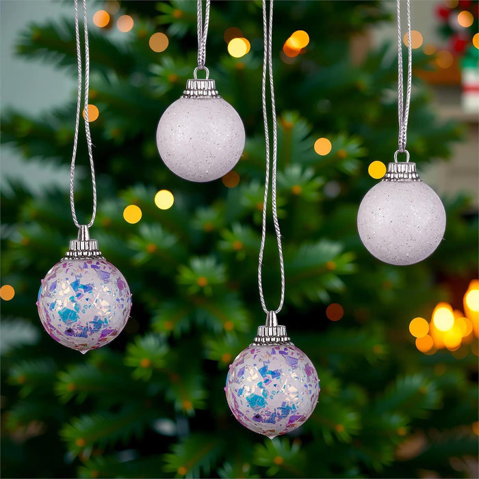 Three decorative hanging Christmas ornaments are shown in front of a blurred Christmas tree with warm lights. Two ornaments are matte light lavender-gray, and one is translucent lavender with blue and purple glitter.