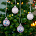 Three decorative hanging Christmas ornaments are shown in front of a blurred Christmas tree with warm lights. Two ornaments are matte light lavender-gray, and one is translucent lavender with blue and purple glitter.