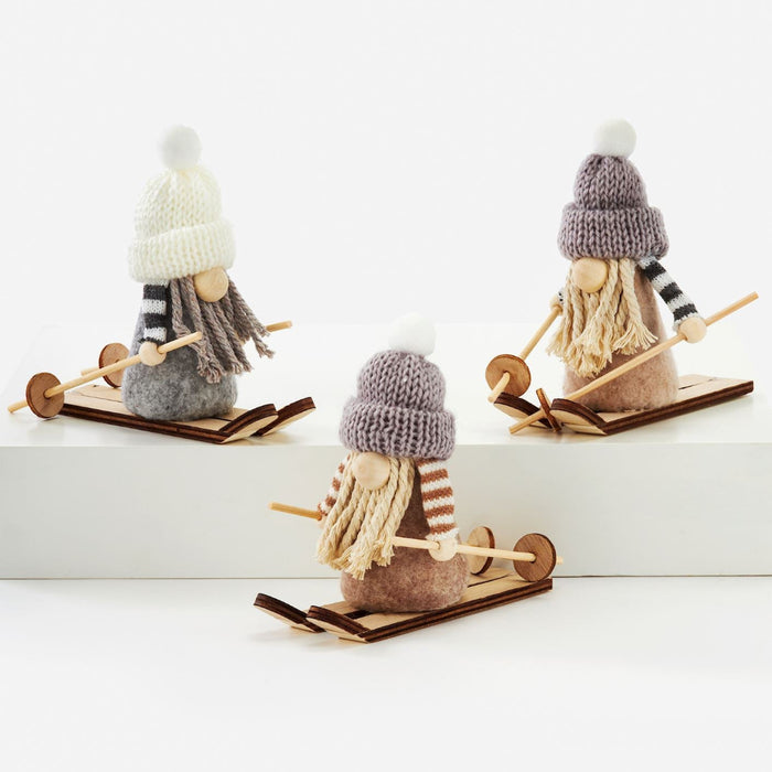 Three small decorative gnome figures wearing knit winter hats and long yarn beards, each mounted on a wooden sled with skis, photographed on a clean white studio background.