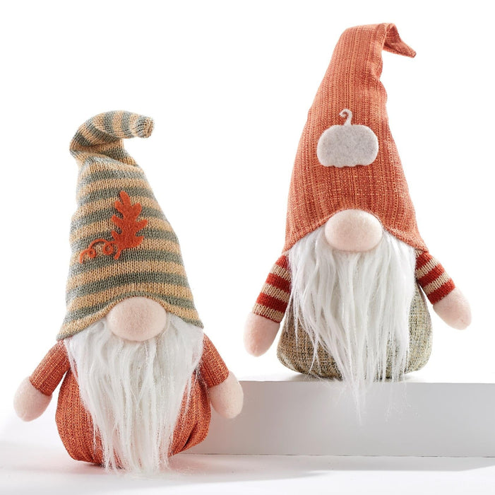 Two knitted autumn gnome dolls with long white beards, one wearing a striped tan and orange hat with a small orange leaf, and the other wearing a solid orange hat with a white pumpkin patch, posed on a white riser against a white background.