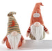 Two knitted autumn gnome dolls with long white beards, one wearing a striped tan and orange hat with a small orange leaf, and the other wearing a solid orange hat with a white pumpkin patch, posed on a white riser against a white background.